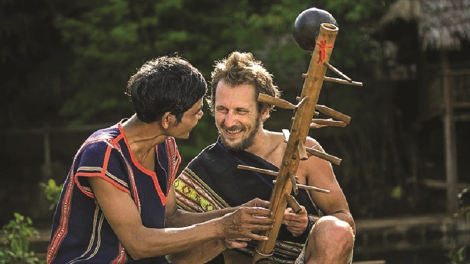 Nghệ nhân Rơ Châm Til (bên trái) đang hướng dẫn một du khách nước ngoài sử dụng nhạc cụ truyền thống Jrai