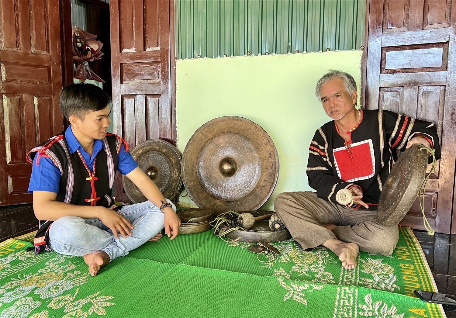 Anh Siu Thanh (bên trái) lắng nghe, học cách đánh cồng chiêng sao cho tiếng vang, phân biệt được các cung bậc âm thanh khác nhau… từ người cha Nay Tek